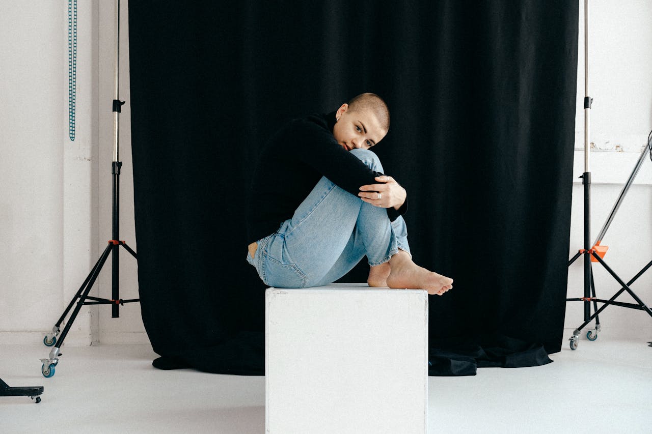 A woman in casual attire sits on a white cube in a professional photo studio with a black backdrop.